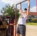 A muscular man in a white sleeveless shirt, black cap, and blue sunglasses performs pull-ups on a red metal pull-up bar station outdoors. A man wearing a black t-shirt and tan tactical/plate carrier vest with a 'SAR' patch observes and steadies the equipment. The setting appears to be an outdoor public event or fair, with a building, trees, and a flag visible in the background.