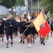 A large formation of U.S. Army soldiers in black PT (physical training) uniforms are conducting a group run, likely a unit formation run or Army birthday run. A soldier in the front carries a large yellow and red unit guidon/flag with a military insignia. The run appears to take place on a military installation with buildings and trees visible in the background.