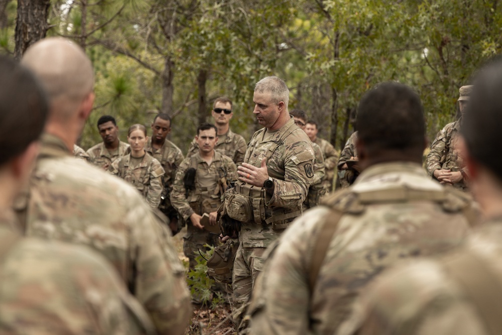 A military leader in camouflage uniform addresses a group of soldiers gathered in a wooded outdoor setting. The central figure appears to be briefing or debriefing the assembled personnel, who are attentively listening. The setting suggests a field training exercise or operational briefing in a forested environment.