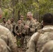 A military leader in camouflage uniform addresses a group of soldiers gathered in a wooded outdoor setting. The central figure appears to be briefing or debriefing the assembled personnel, who are attentively listening. The setting suggests a field training exercise or operational briefing in a forested environment.