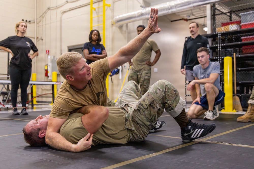 A military combatives training session is taking place on a padded mat inside what appears to be a gymnasium or training facility. One service member in camouflage uniform is mounted on top of another who is on the ground, demonstrating a ground control technique with an arm raised. Several observers in military and civilian attire watch the demonstration from the perimeter.