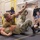 145th AW wing staff participates in combatives A military combatives training session is taking place on a padded mat inside what appears to be a gymnasium or training facility. One service member in camouflage uniform is mounted on top of another who is on the ground, demonstrating a ground control technique with an arm raised. Several observers in military and civilian attire watch the demonstration from the perimeter.