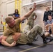 A military combatives training session is taking place on a padded mat inside what appears to be a gymnasium or training facility. One service member in camouflage uniform is mounted on top of another who is on the ground, demonstrating a ground control technique with an arm raised. Several observers in military and civilian attire watch the demonstration from the perimeter.