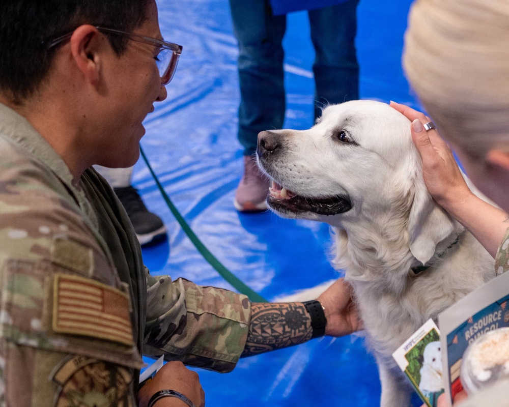 A U.S. military service member in Army/Air Force multicam camouflage uniform with an American flag patch interacts with a white Golden Retriever or similar breed therapy dog at what appears to be a wellness or resource event. The soldier, who has visible tattoos on his forearm and wears glasses, is petting the dog alongside another person. A pamphlet or brochure labeled 'RESOURCE' is visible in the lower right corner, suggesting this is a mental health or support services event.