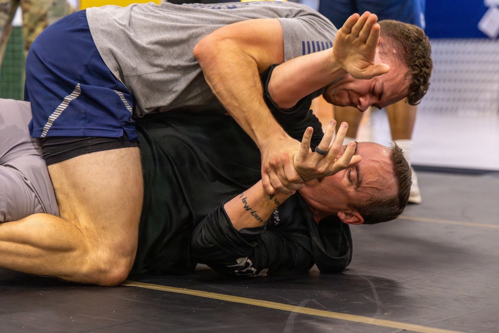 Two athletes are engaged in a ground grappling match, likely Brazilian Jiu-Jitsu or submission wrestling, on a black mat. The top competitor in a gray shirt and blue shorts is applying a face push or wrist control technique against the bottom competitor in a black rash guard. The image captures an intense moment of close-quarters ground fighting with visible physical strain on both athletes.