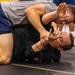 Two athletes are engaged in a ground grappling match, likely Brazilian Jiu-Jitsu or submission wrestling, on a black mat. The top competitor in a gray shirt and blue shorts is applying a face push or wrist control technique against the bottom competitor in a black rash guard. The image captures an intense moment of close-quarters ground fighting with visible physical strain on both athletes.