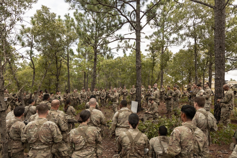 A large group of military personnel in camouflage uniforms are gathered in a wooded area for what appears to be an outdoor briefing or training exercise. A central figure appears to be addressing the assembled soldiers, with a whiteboard or flip chart visible nearby. The setting suggests a field training environment at a military installation.
