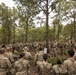 A large group of military personnel in camouflage uniforms are gathered in a wooded area for what appears to be an outdoor briefing or training exercise. A central figure appears to be addressing the assembled soldiers, with a whiteboard or flip chart visible nearby. The setting suggests a field training environment at a military installation.