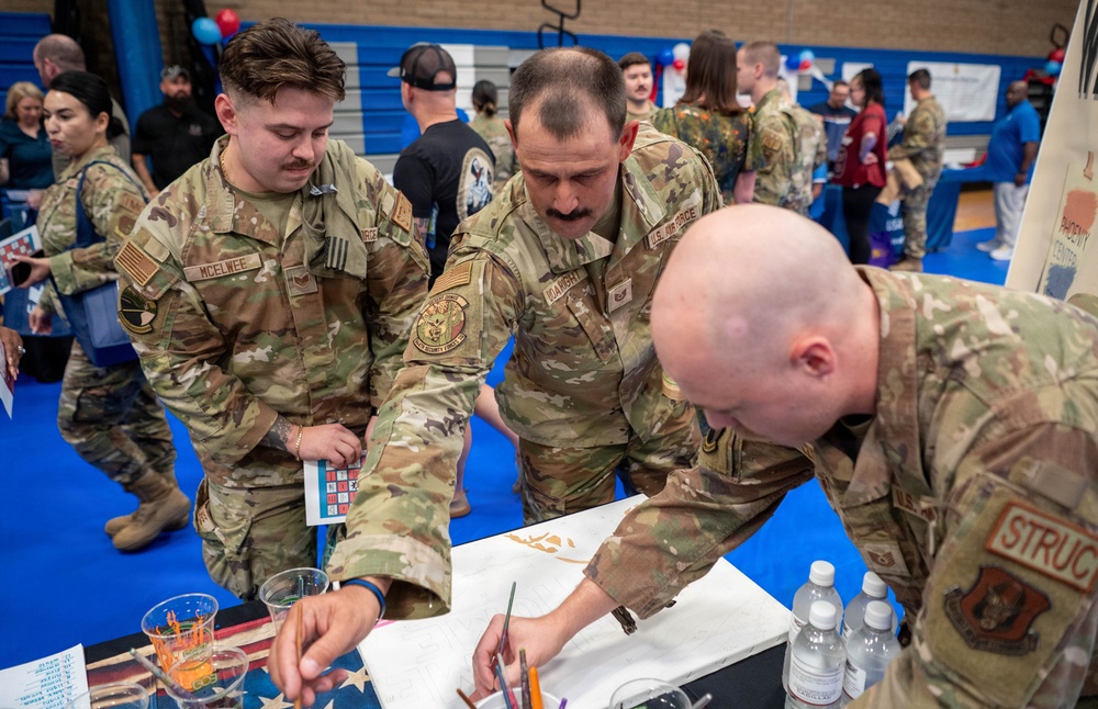 Three U.S. Air Force personnel in OCP (Operational Camouflage Pattern) uniforms are gathered around a table at what appears to be a military community event or resource fair held in a gymnasium. The soldiers appear to be engaged in a painting or art activity at a booth. The background shows additional attendees and display booths decorated with red, white, and blue colors.