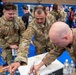 Three U.S. Air Force personnel in OCP (Operational Camouflage Pattern) uniforms are gathered around a table at what appears to be a military community event or resource fair held in a gymnasium. The soldiers appear to be engaged in a painting or art activity at a booth. The background shows additional attendees and display booths decorated with red, white, and blue colors.