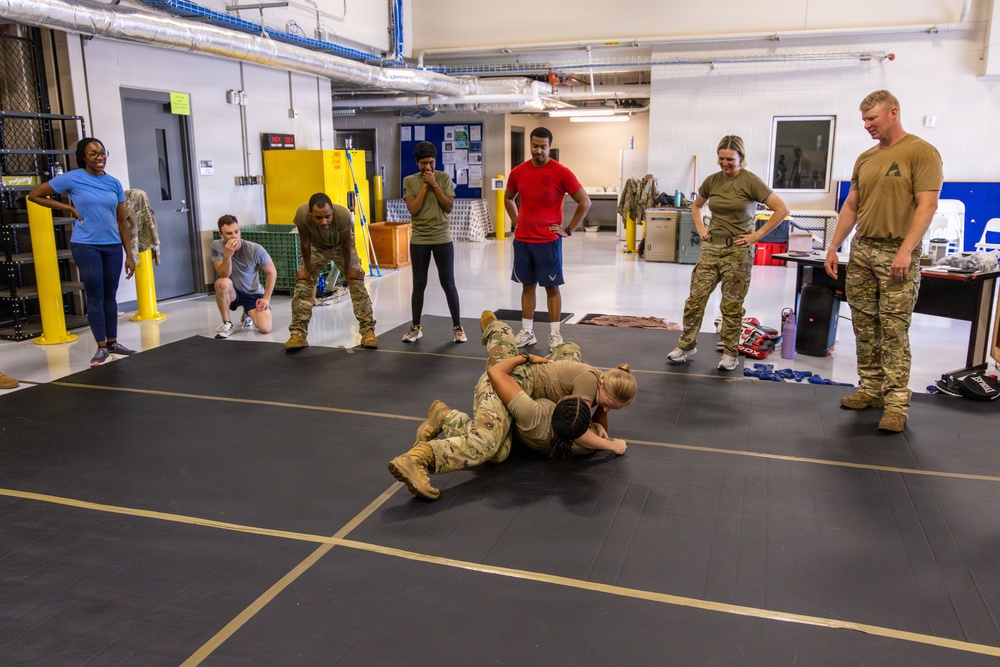 A group of military personnel and civilians are gathered in an indoor training facility watching two individuals in camouflage uniforms practicing ground combat or combatives techniques on a black mat. Observers are standing and kneeling around the perimeter of the mat in a semicircle, some in military uniforms and others in civilian athletic wear. The setting appears to be a military training room or gymnasium with industrial features.