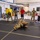 145th AW wing staff participates in combatives A group of military personnel and civilians are gathered in an indoor training facility watching two individuals in camouflage uniforms practicing ground combat or combatives techniques on a black mat. Observers are standing and kneeling around the perimeter of the mat in a semicircle, some in military uniforms and others in civilian athletic wear. The setting appears to be a military training room or gymnasium with industrial features.