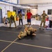 A group of military personnel and civilians are gathered in an indoor training facility watching two individuals in camouflage uniforms practicing ground combat or combatives techniques on a black mat. Observers are standing and kneeling around the perimeter of the mat in a semicircle, some in military uniforms and others in civilian athletic wear. The setting appears to be a military training room or gymnasium with industrial features.