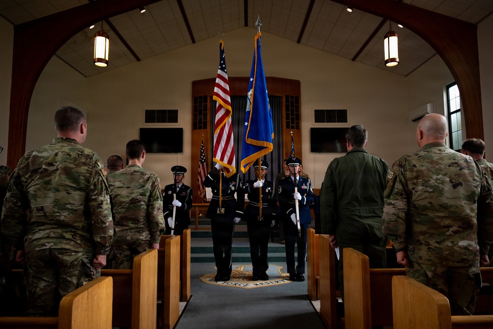 A military color guard ceremony is taking place inside what appears to be a chapel on a U.S. Air Force Reserve base, as indicated by the seal visible on the floor. Members of the color guard in dress blues are presenting the U.S. flag and Air Force flag while uniformed personnel in camouflage and flight suits stand at attention in the pews. The formal setting and attire suggest an official military ceremony or memorial service.