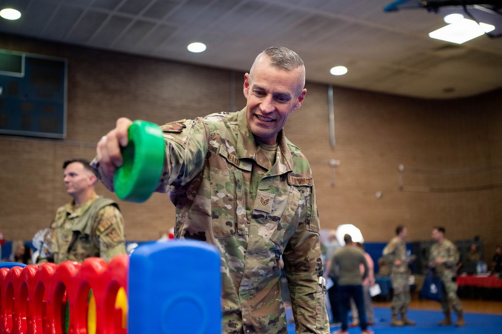A U.S. Air Force staff sergeant in OCP (Operational Camouflage Pattern) uniform is smiling while engaging with colorful children's toys, likely at a family or community event held in a gymnasium or recreation facility on a military installation. Several other uniformed personnel are visible in the background, suggesting an organized military community event. The atmosphere appears relaxed and informal.
