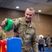 A U.S. Air Force staff sergeant in OCP (Operational Camouflage Pattern) uniform is smiling while engaging with colorful children's toys, likely at a family or community event held in a gymnasium or recreation facility on a military installation. Several other uniformed personnel are visible in the background, suggesting an organized military community event. The atmosphere appears relaxed and informal.