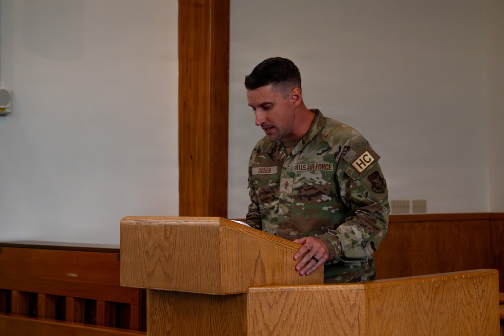 A U.S. Air Force service member identified by name tape as 'OGDEN' stands at a wooden podium, appearing to read or speak from documents. The individual wears Operational Camouflage Pattern (OCP) uniform with 'U.S. AIR FORCE' tape, 'HC' (Chaplain Corps) shoulder patch, and a chaplain cross insignia on the chest. The setting appears to be a chapel or formal meeting room with wooden furnishings.
