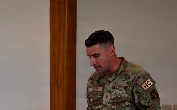 A U.S. Air Force service member identified by name tape as 'OGDEN' stands at a wooden podium, appearing to read or speak from documents. The individual wears Operational Camouflage Pattern (OCP) uniform with 'U.S. AIR FORCE' tape, 'HC' (Chaplain Corps) shoulder patch, and a chaplain cross insignia on the chest. The setting appears to be a chapel or formal meeting room with wooden furnishings.