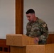 A U.S. Air Force service member identified by name tape as 'OGDEN' stands at a wooden podium, appearing to read or speak from documents. The individual wears Operational Camouflage Pattern (OCP) uniform with 'U.S. AIR FORCE' tape, 'HC' (Chaplain Corps) shoulder patch, and a chaplain cross insignia on the chest. The setting appears to be a chapel or formal meeting room with wooden furnishings.