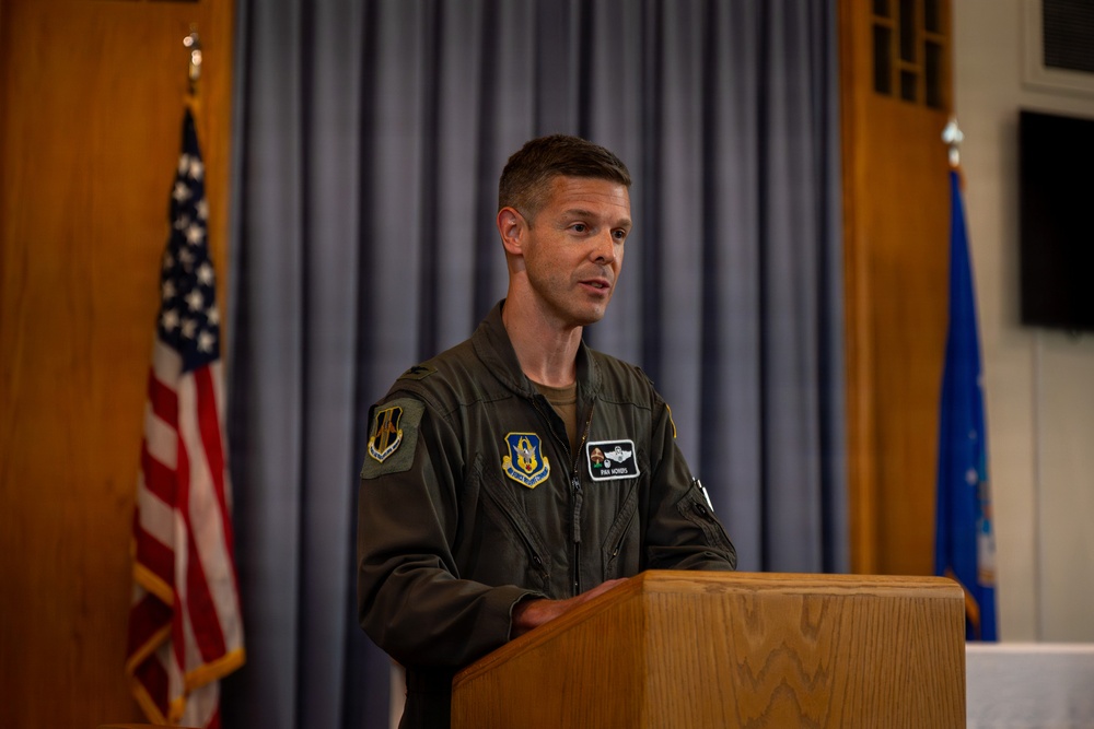 A U.S. Air Force officer in an olive green flight suit is speaking at a wooden podium. He is positioned in front of a gray curtain backdrop with a U.S. flag to his left and a U.S. Air Force flag to his right. The setting appears to be an official military ceremony or briefing.