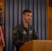 A U.S. Air Force officer in an olive green flight suit is speaking at a wooden podium. He is positioned in front of a gray curtain backdrop with a U.S. flag to his left and a U.S. Air Force flag to his right. The setting appears to be an official military ceremony or briefing.