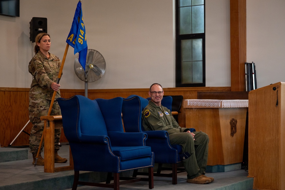 A military ceremony taking place in what appears to be a chapel or auditorium setting. A female soldier in camouflage uniform stands holding a blue unit guidon flag while a male officer in an olive green flight suit sits in a large blue wingback chair. The setting and arrangement suggest a formal change of command or unit activation/deactivation ceremony.