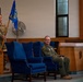 A military ceremony taking place in what appears to be a chapel or auditorium setting. A female soldier in camouflage uniform stands holding a blue unit guidon flag while a male officer in an olive green flight suit sits in a large blue wingback chair. The setting and arrangement suggest a formal change of command or unit activation/deactivation ceremony.