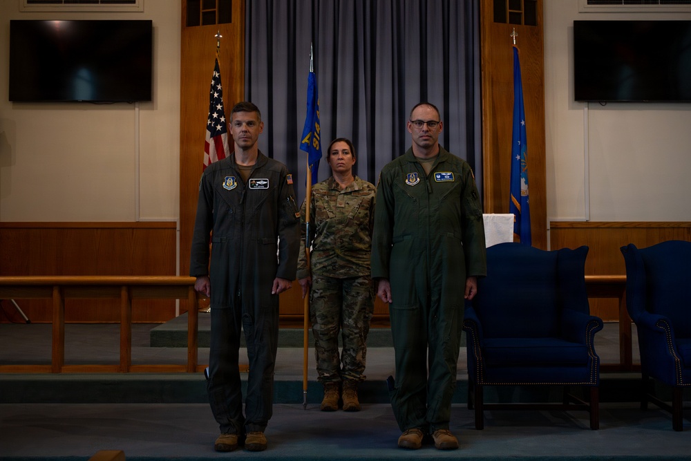 Three military personnel stand at attention in a formal indoor ceremony setting. Two individuals wear olive green flight suits with Air Force patches, while one individual in the center wears OCP (Operational Camouflage Pattern) combat uniform and holds a unit guidon flag. The setting appears to be a military chapel or ceremonial hall, suggesting a change of command or similar formal military ceremony.