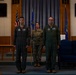 Three military personnel stand at attention in a formal indoor ceremony setting. Two individuals wear olive green flight suits with Air Force patches, while one individual in the center wears OCP (Operational Camouflage Pattern) combat uniform and holds a unit guidon flag. The setting appears to be a military chapel or ceremonial hall, suggesting a change of command or similar formal military ceremony.