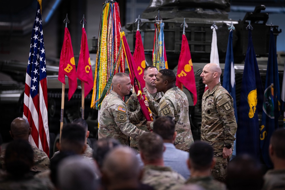 A U.S. Army military ceremony, likely a change of command or activation/deactivation ceremony, is taking place in what appears to be a hangar or large indoor facility. Two soldiers in the foreground are exchanging or passing a unit guidon (flag), which is the central symbolic act of such ceremonies. An audience of uniformed personnel and civilians observes the proceedings.