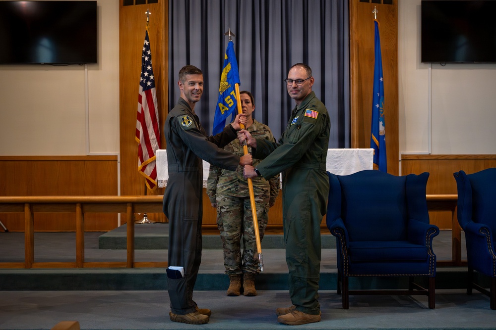 A military change of command or activation ceremony is taking place in what appears to be a chapel or formal ceremony room on a U.S. Air Force installation. Two personnel in flight suits are exchanging a guidon (unit flag on a staff), while a third service member in camouflage uniform serves as the guidon bearer. This type of ceremony formally transfers command authority from one officer to another.