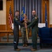 A military change of command or activation ceremony is taking place in what appears to be a chapel or formal ceremony room on a U.S. Air Force installation. Two personnel in flight suits are exchanging a guidon (unit flag on a staff), while a third service member in camouflage uniform serves as the guidon bearer. This type of ceremony formally transfers command authority from one officer to another.