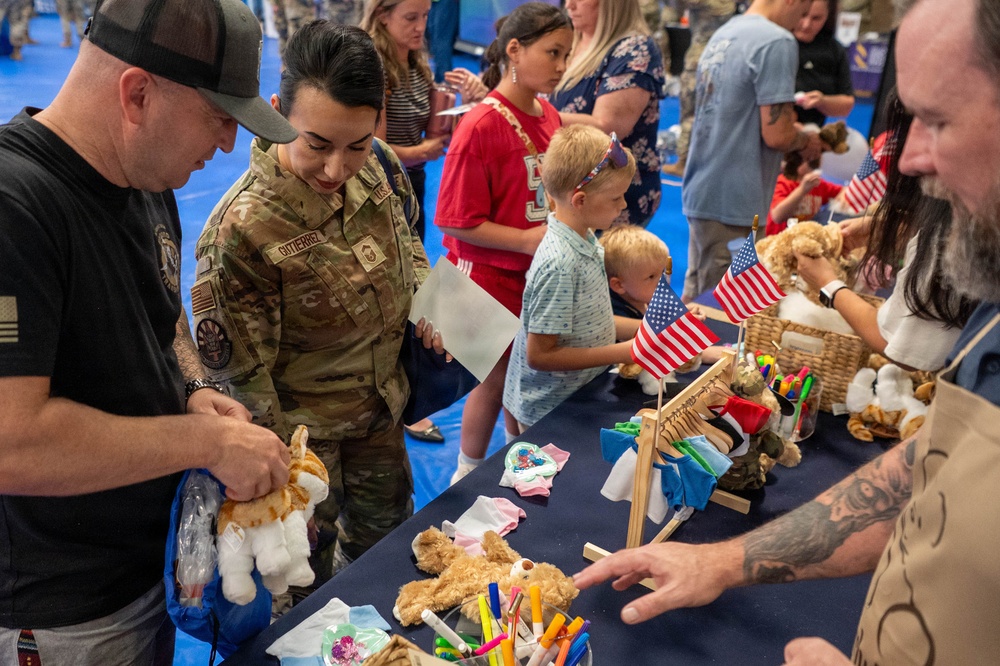 A military family event featuring a stuffed animal building station, similar to a Build-A-Bear activity. A uniformed service member with the name tape 'GUTIERREZ' in camouflage uniform participates alongside civilians, children, and other attendees. American flags and small stuffed animals are displayed on a table covered with a dark blue cloth.