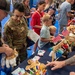 A military family event featuring a stuffed animal building station, similar to a Build-A-Bear activity. A uniformed service member with the name tape 'GUTIERREZ' in camouflage uniform participates alongside civilians, children, and other attendees. American flags and small stuffed animals are displayed on a table covered with a dark blue cloth.