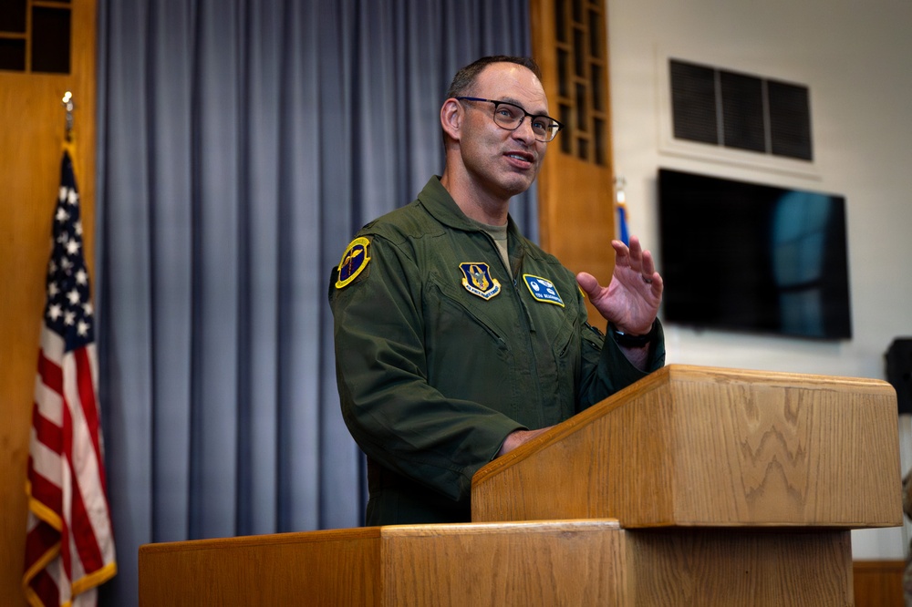 A military officer in a green flight suit stands at a wooden podium delivering a speech or briefing. He is wearing glasses and gesturing with his right hand, appearing engaged in his address. The setting appears to be an official military briefing room with an American flag and blue curtains visible in the background.