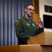 A military officer in a green flight suit stands at a wooden podium delivering a speech or briefing. He is wearing glasses and gesturing with his right hand, appearing engaged in his address. The setting appears to be an official military briefing room with an American flag and blue curtains visible in the background.