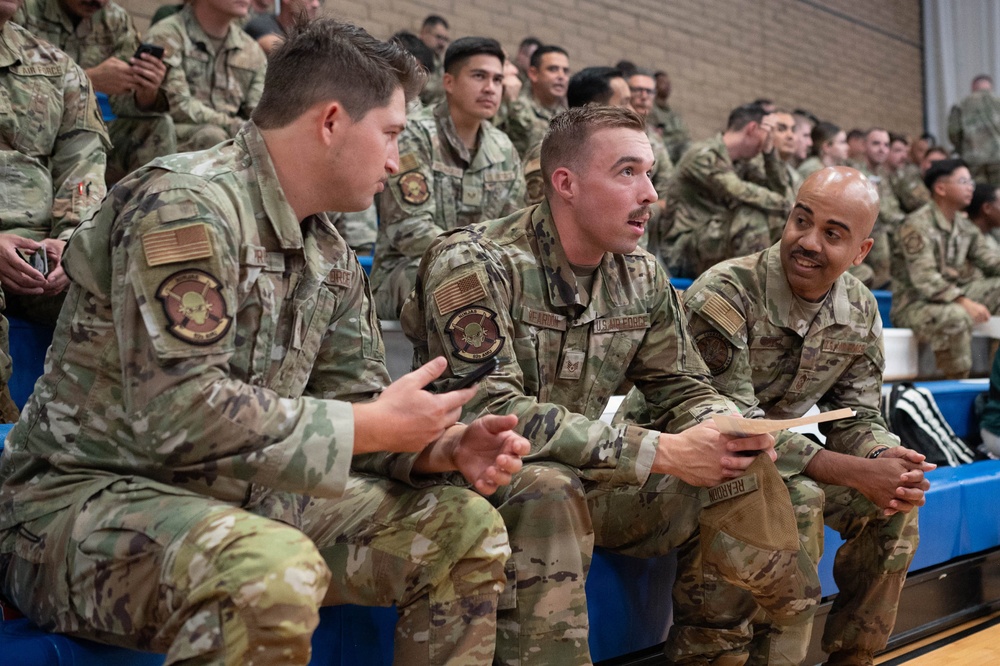 A group of U.S. Air Force personnel in OCP (Operational Camouflage Pattern) uniforms are seated in bleachers, likely inside a gymnasium or indoor facility. Three airmen in the foreground appear to be engaged in an animated conversation, with one holding what appears to be a small piece of paper or card. A large formation of additional uniformed personnel fills the bleachers in the background.