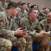A group of U.S. Air Force personnel in OCP (Operational Camouflage Pattern) uniforms are seated in bleachers, likely inside a gymnasium or indoor facility. Three airmen in the foreground appear to be engaged in an animated conversation, with one holding what appears to be a small piece of paper or card. A large formation of additional uniformed personnel fills the bleachers in the background.