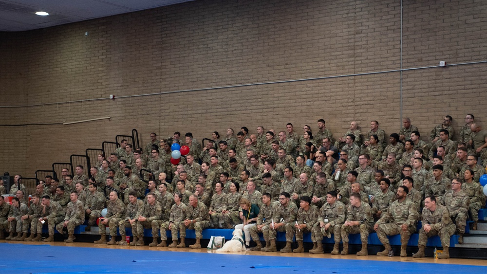 A large group of military personnel in camouflage uniforms are seated in bleachers inside what appears to be a gymnasium or indoor sports facility. The gathering appears to be a formal military assembly or ceremony, indicated by the organized seating arrangement and the presence of red, white, and blue balloons. A service dog and its handler are visible seated at the front row among the uniformed personnel.