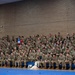 A large group of military personnel in camouflage uniforms are seated in bleachers inside what appears to be a gymnasium or indoor sports facility. The gathering appears to be a formal military assembly or ceremony, indicated by the organized seating arrangement and the presence of red, white, and blue balloons. A service dog and its handler are visible seated at the front row among the uniformed personnel.