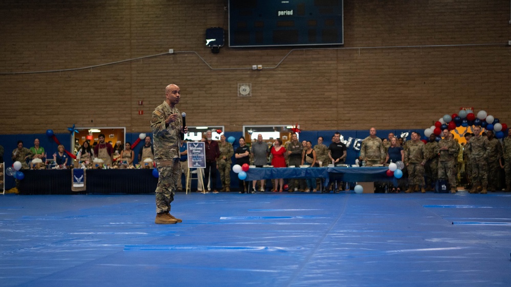 A military officer in camouflage uniform stands on a blue gymnasium floor holding a microphone, addressing a large crowd of military personnel and civilians gathered along the perimeter. The event appears to be a formal military ceremony or welcome home celebration, indicated by red, white, and blue balloon decorations and banners visible in the background. The setting is an indoor gymnasium or sports facility on what appears to be a military installation.