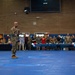 A military officer in camouflage uniform stands on a blue gymnasium floor holding a microphone, addressing a large crowd of military personnel and civilians gathered along the perimeter. The event appears to be a formal military ceremony or welcome home celebration, indicated by red, white, and blue balloon decorations and banners visible in the background. The setting is an indoor gymnasium or sports facility on what appears to be a military installation.