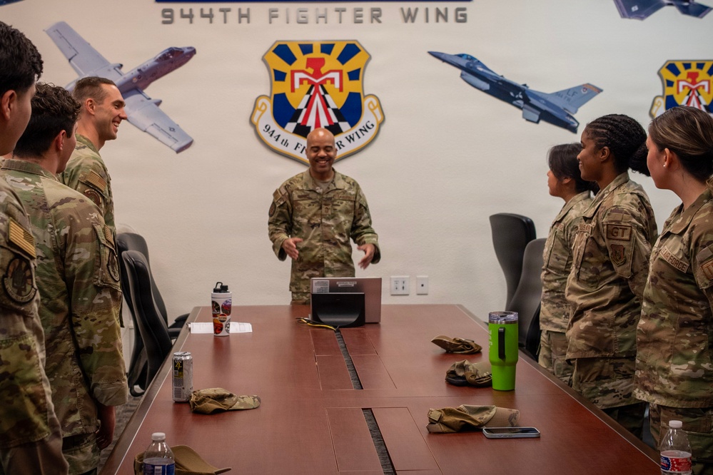 A group of U.S. Air Force personnel in OCP (Operational Camouflage Pattern) uniforms are gathered around a conference table at the 944th Fighter Wing. A senior airman or officer stands at the head of the table, appearing to address or brief the group in an informal, engaged manner. The setting is an official briefing or meeting room decorated with the 944th Fighter Wing emblem and aircraft imagery.