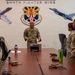 A group of U.S. Air Force personnel in OCP (Operational Camouflage Pattern) uniforms are gathered around a conference table at the 944th Fighter Wing. A senior airman or officer stands at the head of the table, appearing to address or brief the group in an informal, engaged manner. The setting is an official briefing or meeting room decorated with the 944th Fighter Wing emblem and aircraft imagery.