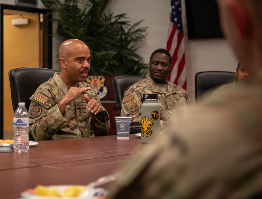 Military personnel in OCP (Operational Camouflage Pattern) uniforms are seated around a conference table during what appears to be a briefing or discussion session. The primary speaker, a bald male service member, is gesturing with his hands while addressing others at the table. The setting appears to be a formal military conference room with a U.S. flag and unit insignia visible in the background.
