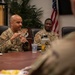 Military personnel in OCP (Operational Camouflage Pattern) uniforms are seated around a conference table during what appears to be a briefing or discussion session. The primary speaker, a bald male service member, is gesturing with his hands while addressing others at the table. The setting appears to be a formal military conference room with a U.S. flag and unit insignia visible in the background.