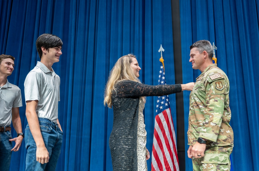 A military promotion or award ceremony is taking place on a stage with a blue curtain backdrop and an American flag. A woman is pinning an insignia or badge onto the uniform of a male U.S. Air Force service member in OCP (Operational Camouflage Pattern) uniform, while two young men look on smiling. The setting and participants suggest this is a family pinning ceremony, likely marking a promotion in rank.