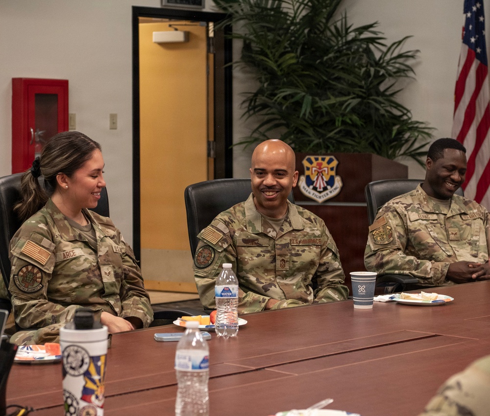 Three U.S. Air Force personnel in OCP (Operational Camouflage Pattern) uniforms are seated at a conference table, smiling and engaged in conversation. The setting appears to be an official military briefing or meeting room, identifiable by the unit crest on the wall and the U.S. flag. The atmosphere is relaxed and informal, with food and beverages on the table.