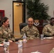 Three U.S. Air Force personnel in OCP (Operational Camouflage Pattern) uniforms are seated at a conference table, smiling and engaged in conversation. The setting appears to be an official military briefing or meeting room, identifiable by the unit crest on the wall and the U.S. flag. The atmosphere is relaxed and informal, with food and beverages on the table.