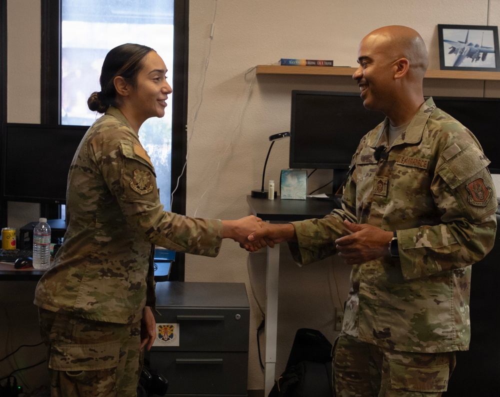 Two U.S. Air Force personnel in OCP (Operational Camouflage Pattern) uniforms are shaking hands in what appears to be an office setting. The male service member on the right has 'U.S. AIR FORCE' tape visible on his uniform and appears to be of senior rank. The interaction appears to be a formal recognition or greeting ceremony.